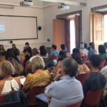 várias pessoas numa plateia assistem a conferência. 5 pessoas numa mesa de comunicação, com ecrã em fundo.