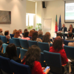 sala com plateia e mesa com quatro pessoas. em fundo, implementar orçamentos sensíveis ao género