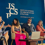 no palco de uma conferência, 5 pessoas sentadas. uma mulher segura um microfone.