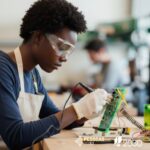 mulher com óculos protetores, solda pequenos componentes eletrónicos numa placa de circuito